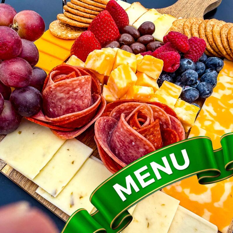 "A decorative, arched green satin ribbon with gold trim and the word 'MENU' in bold white text, layered over a vibrant platter of assorted meats and cheeses."