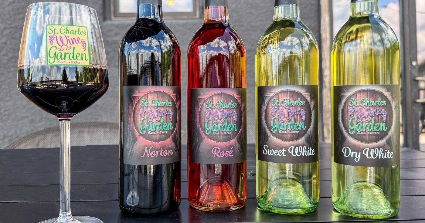 A large wine glass filled with red wine stands next to a lineup of four wine bottles on an outdoor table. The bottles feature custom St. Charles Wine Garden labels showcasing their house varieties from left to right: Norton red wine, Rosé, Sweet White, and Dry White.